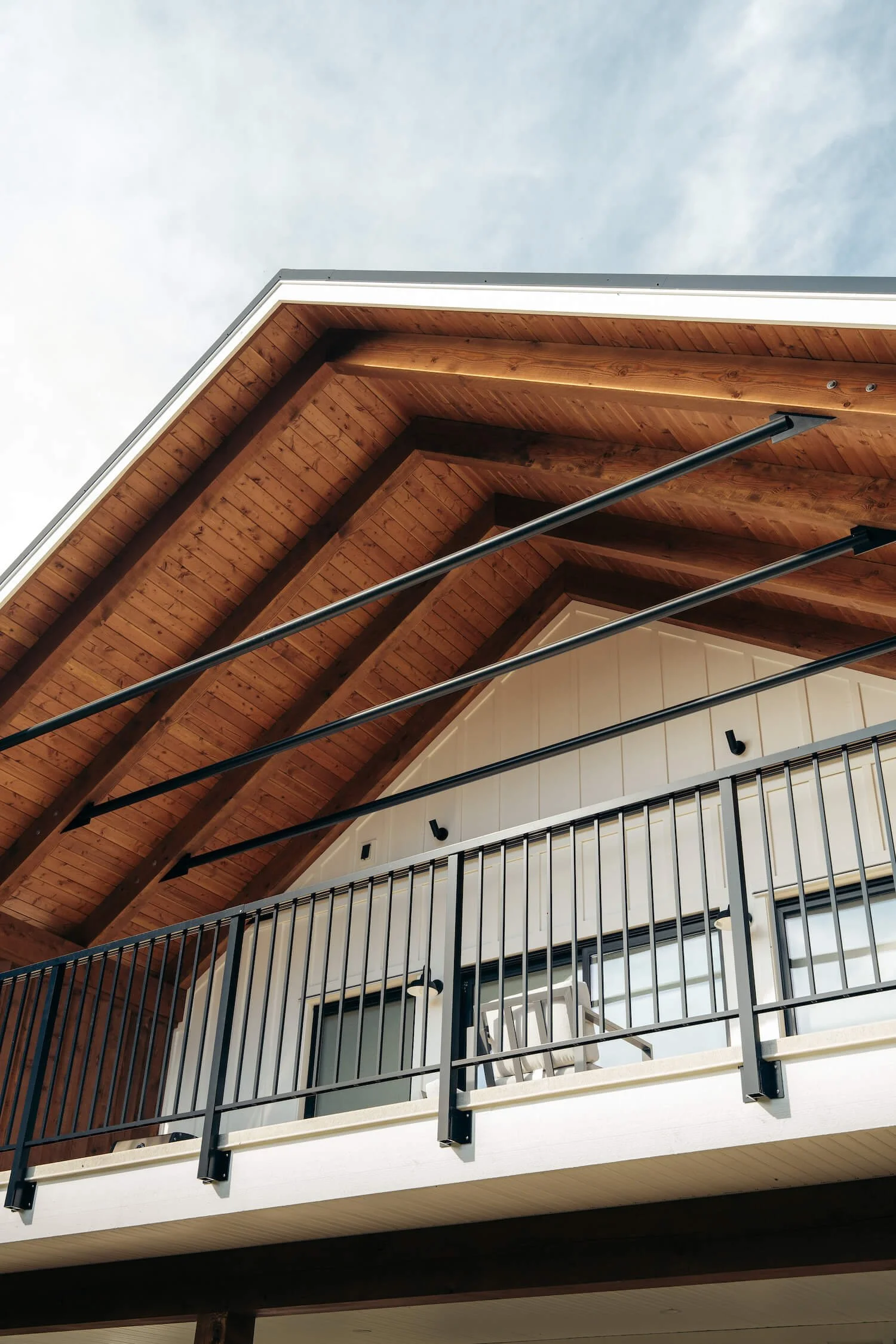 Exterior view of a modern house's balcony with black metal railing, wooden ceiling, and white wall with two black outdoor lights.