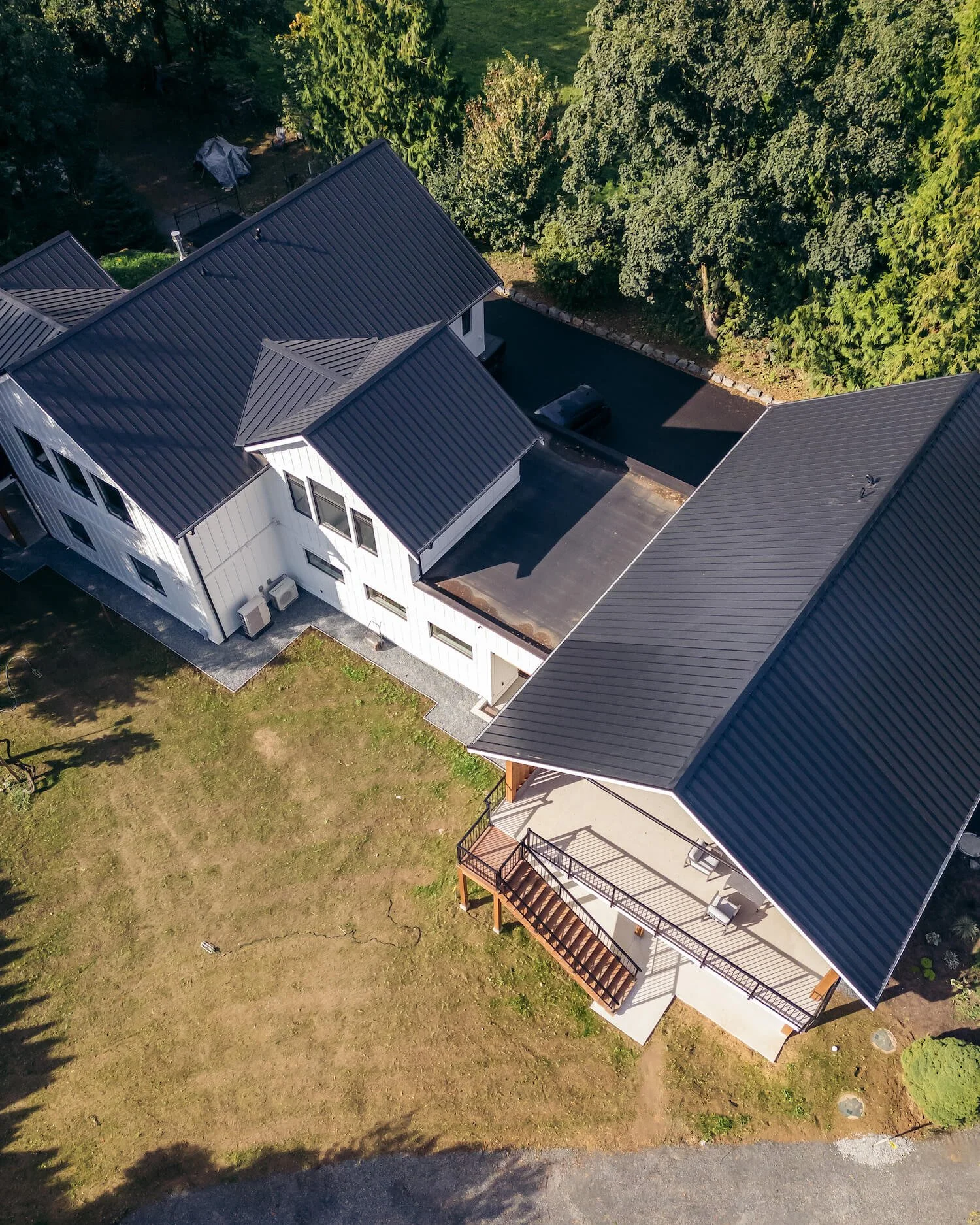 An aerial view of a residential property with multiple buildings, surrounded by trees, a driveway with parked cars, a large green lawn, and farmland.
