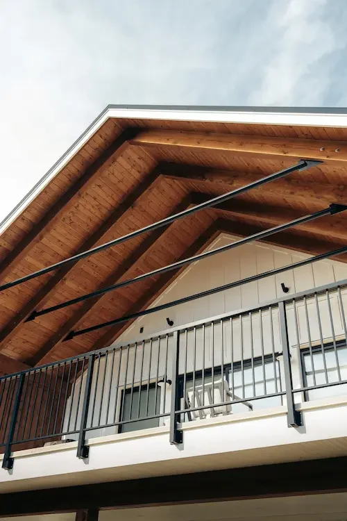 Exterior view of a modern house's balcony with black metal railing, wooden ceiling, and white wall with two black outdoor lights.