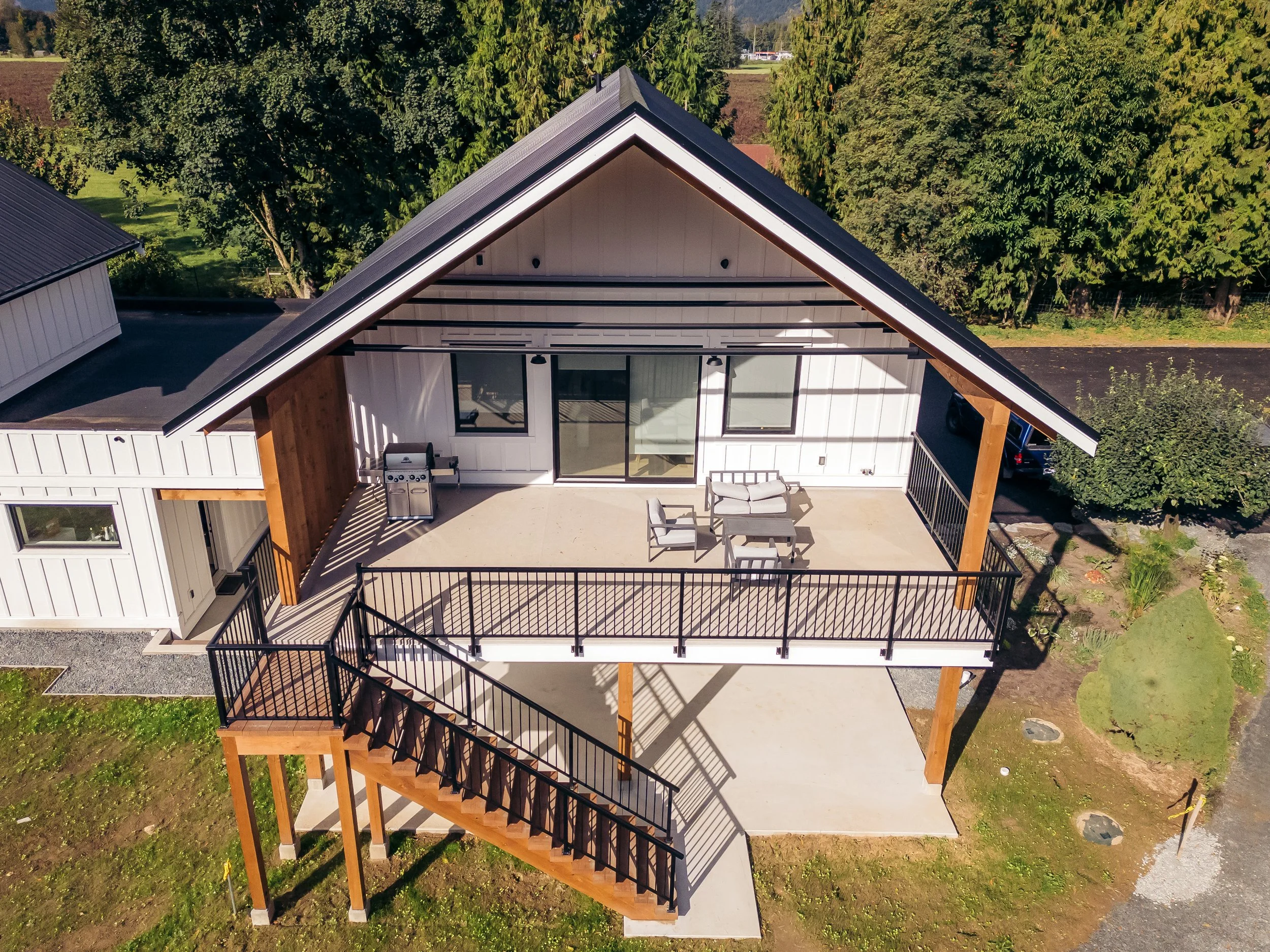 Aerial view of a modern house with a large front porch, black railings, outdoor furniture, and a grill, surrounded by green trees and landscaping.