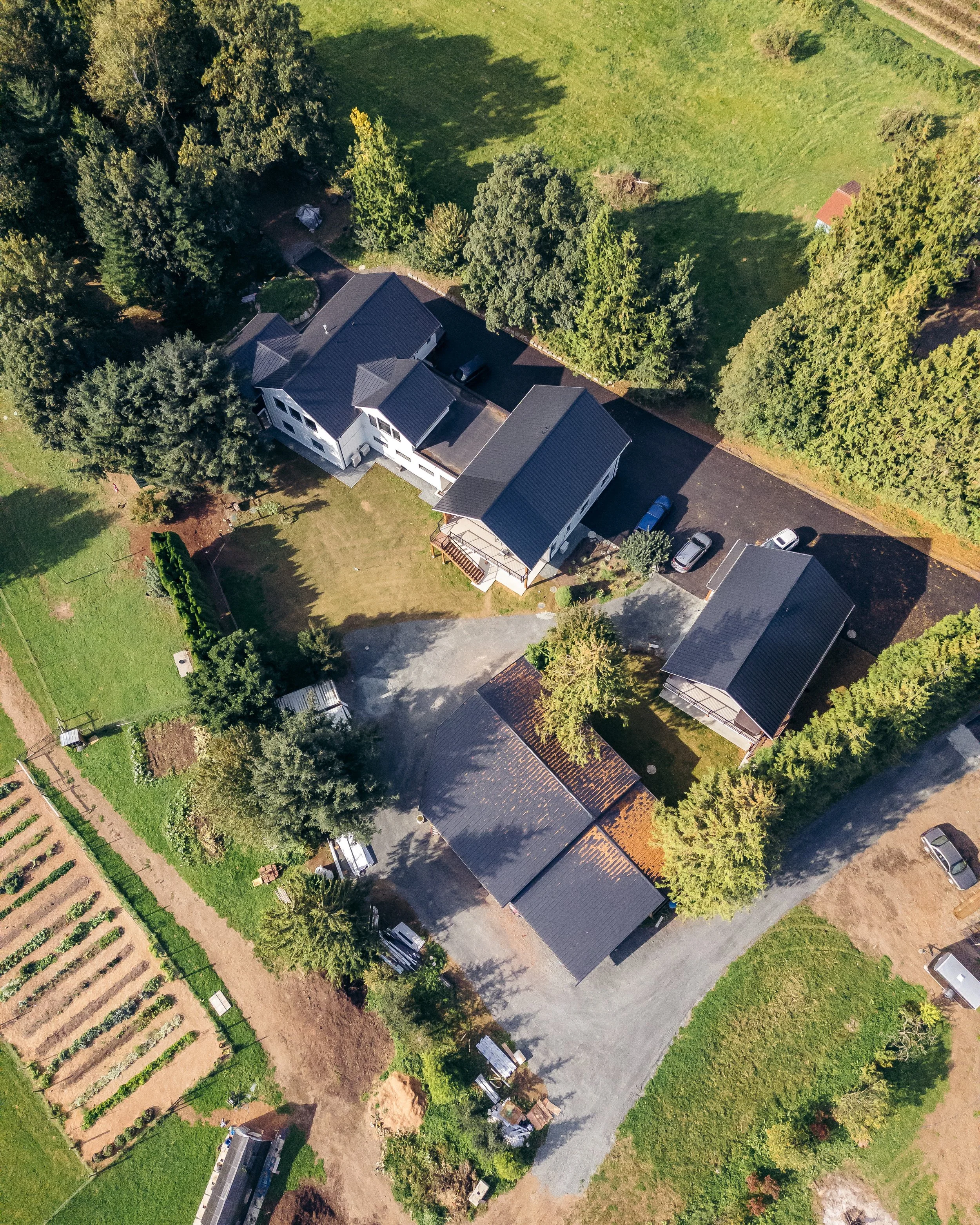 An aerial view of a residential property with multiple buildings, surrounded by trees, a driveway with parked cars, a large green lawn, and farmland.