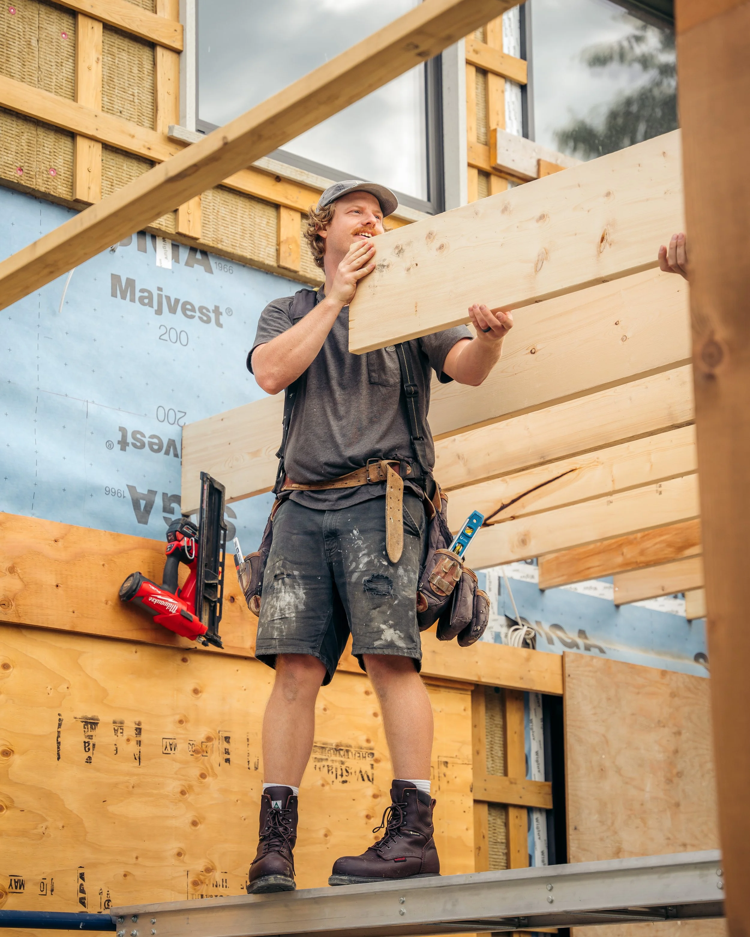 A construction worker in a grey t-shirt, black shorts, and work boots, holding a wooden plank on a building site with wooden framing and blue insulation material behind him.
