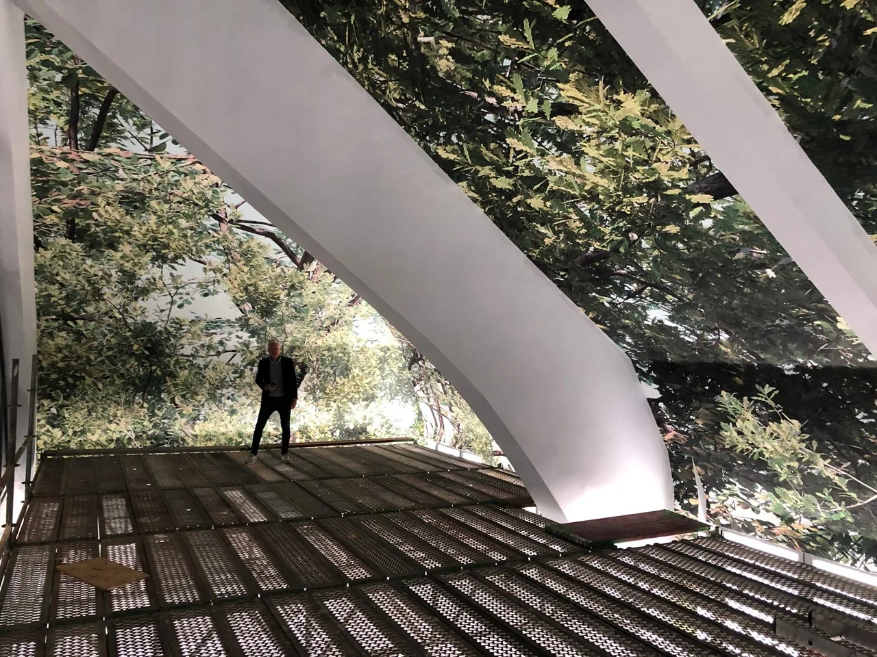 Ein Mann in einem schwarzen Anzug steht auf einer Plattform im Inneren eines Gebäudes mit großen, weißen, bogenförmigen Strukturen und einer Wand mit bepflanztem Außenbereich im Hintergrund.