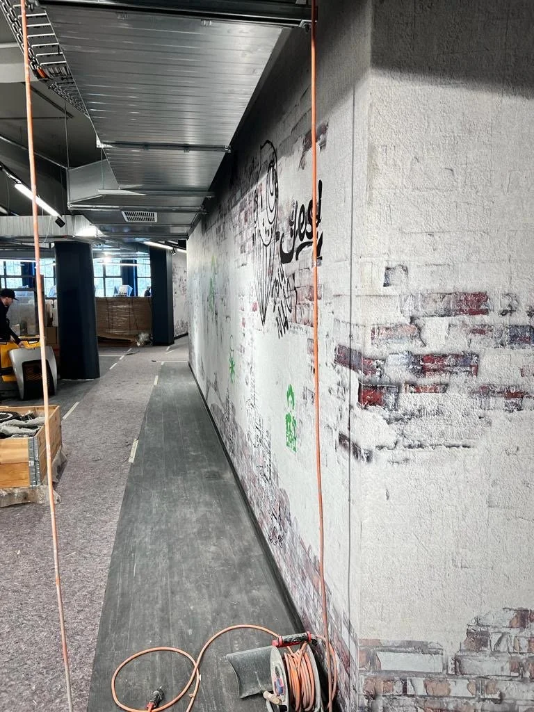 Ein langer Innenraum-Korridor unter Renovierung mit einer weißen Wand, die teilweise mit Graffiti bedeckt ist, und einem Bodenbelag aus dunklem Holz. Es sind Baustoffe und Elektrogeräte im Bereich links zu sehen.