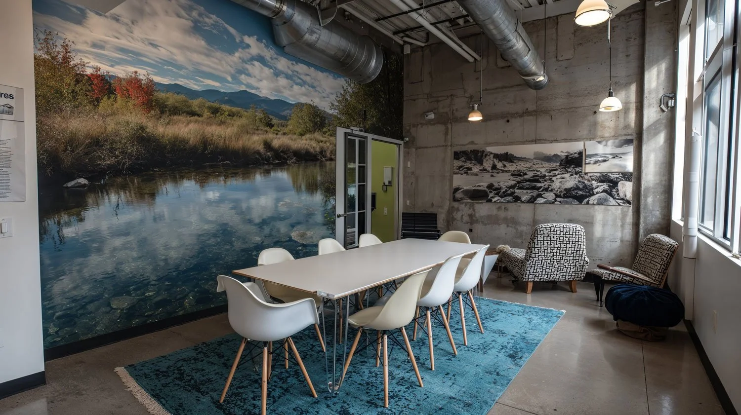 Ein moderner Besprechungsraum mit einem großen weißen Tisch, umgeben von weißen Stühlen, auf einem blauen Teppich. Die Wände sind gestaltet mit Naturfotos, eine zeigt einen Fluss in der Natur, eine andere eine felsige Küstenlandschaft. Es gibt große 