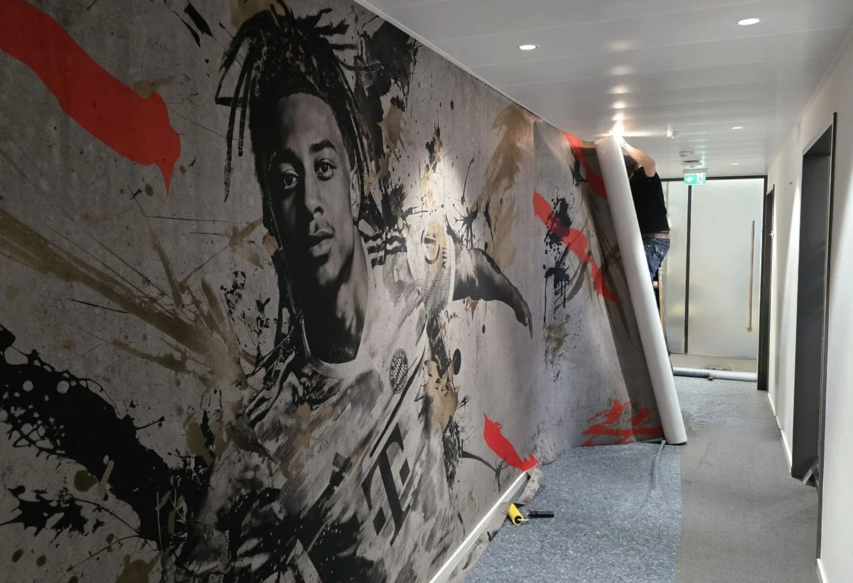 Ein Flur mit einer Wand, auf der ein Wandbild eines berühmten Fußballspielers, wahrscheinlich ein Fußballspieler des FC Bayern München, in Schwarzweiß mit Spritz- und Farbstreifen gestaltet ist. Es wird an der Wand gearbeitet, eine Person installiert eine Wandfolie, die teilweise abrollt.