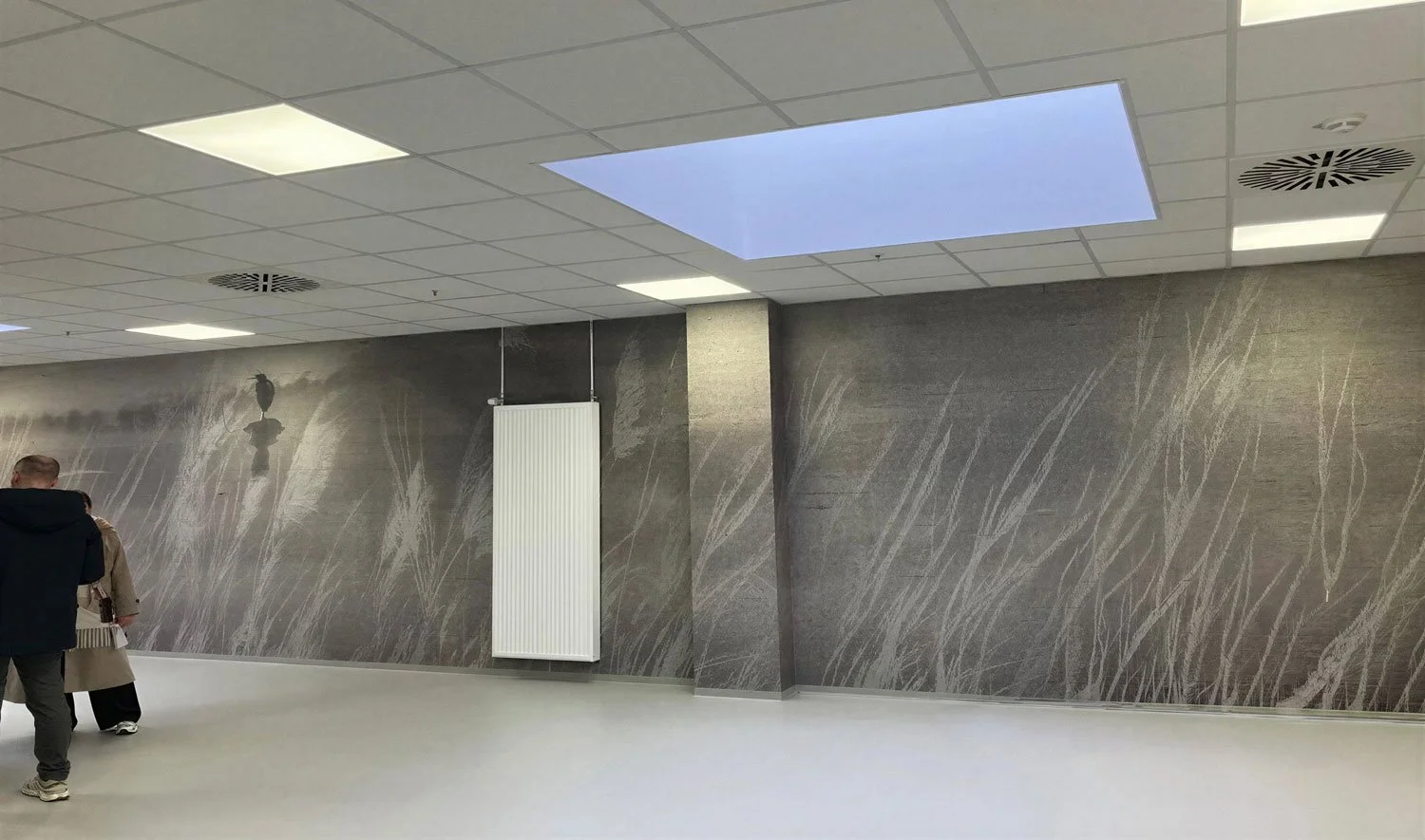 Innenansicht eines modernen Raumes mit grauer Wandgestaltung, einem weißen Heizkörper, einem Deckenfenster, einigen Menschen, die gehen, und einem Deckenventilator.