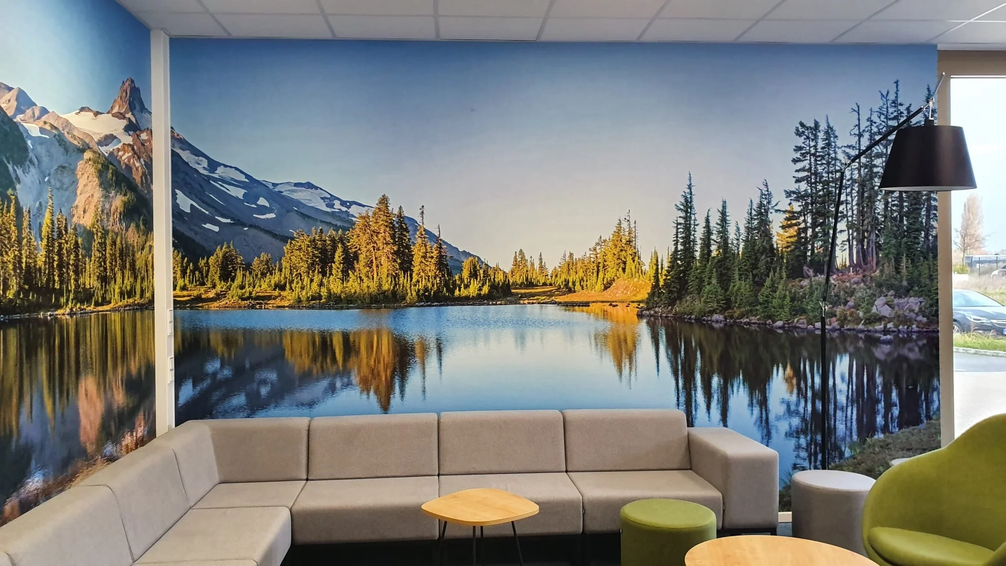 Wand mit Wandbild einer Seen- und Berglandschaft, Wohnzimmer mit grauer Couch, grünem Sessel, kleiner beiger Hocker, runder Holztisch, Stehlampe im Raum.