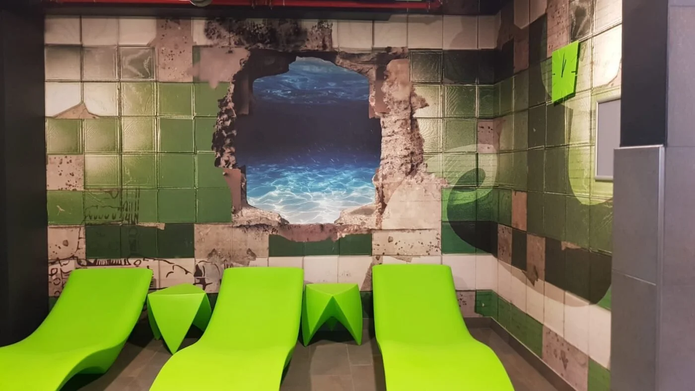 Wartebereich mit grünen Liegesesseln und Wanddekoration, die ein Loch mit einer Wasser- und Meeresansicht zeigt.