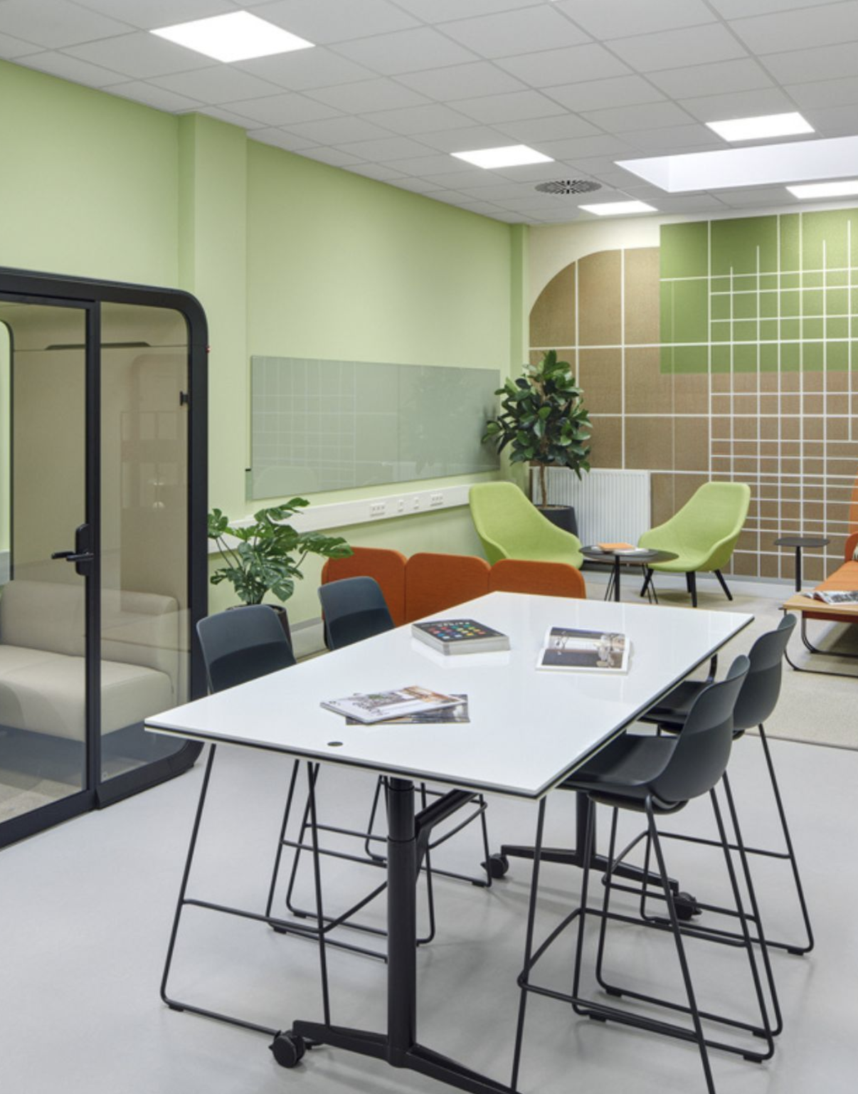 Moderne, helle Meeting- oder Gemeinschaftsraum mit weißen Tischen, schwarzen Stühlen, grünen und orangefarbenen Sitzgelegenheiten, mehreren Pflanzen und geometrischen Wanddekorationen.
