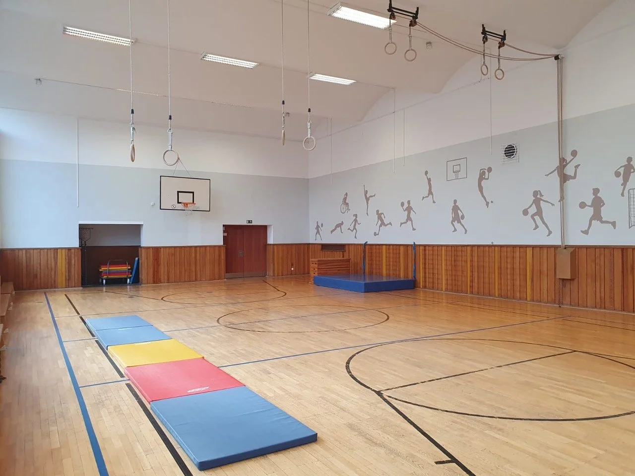 Leerer Turnraum mit Holzfußboden, Turnmatten in verschiedenen Farben, Wandbilder mit Sportsketchen, Basketballkorb, Luftventilator an der Wand, Turngeräte und Gymnastikringe an der Decke.