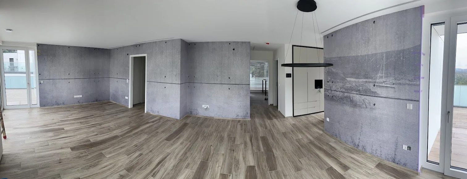 Leeres, modern eingerichtetes Zimmer mit Holzfußboden, grauen Wänden und großen Fenstern/Balkontüren, die einen Balkon zeigen.