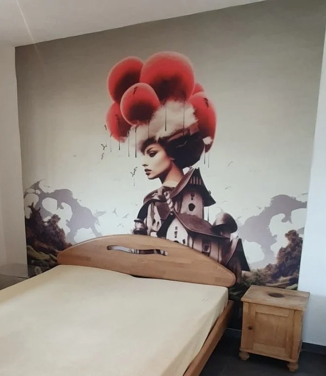 Wandbild mit einem grafischen Porträt einer Frau mit rotem, ballonartigem Haar und windigem Hintergrund. Ein Holzbett und Nachttisch im Vordergrund.