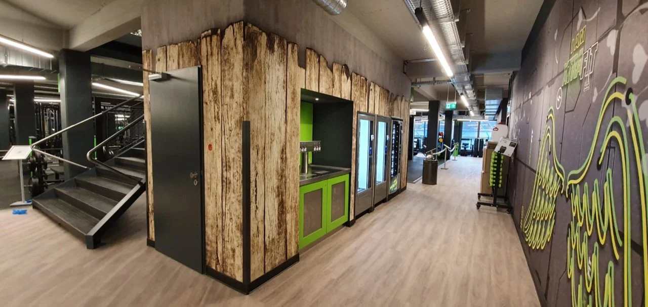 Innenraum eines Fitnessstudios mit Getränkeautomat, Wasserstation, Treppen und Wandkunst.