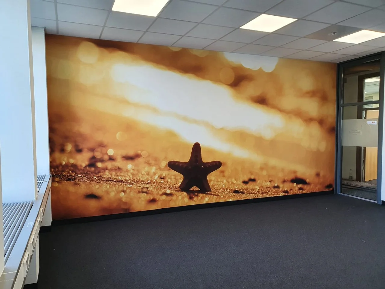 Wand mit einem großen Bild von einem Sandstrand mit einer Sanduhr in Form eines Sterns, aufgenommen bei Sonnenuntergang oder Sonnenaufgang.