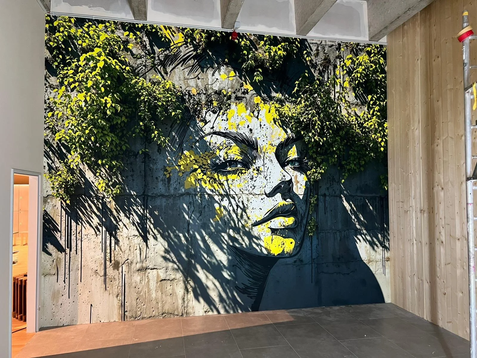Streetart einer Frau mit grünen Blättern im Haar auf einer grauen Wand, mit Schattenwurf und Farbklecksen in Gelb, Schwarz und Weiß.