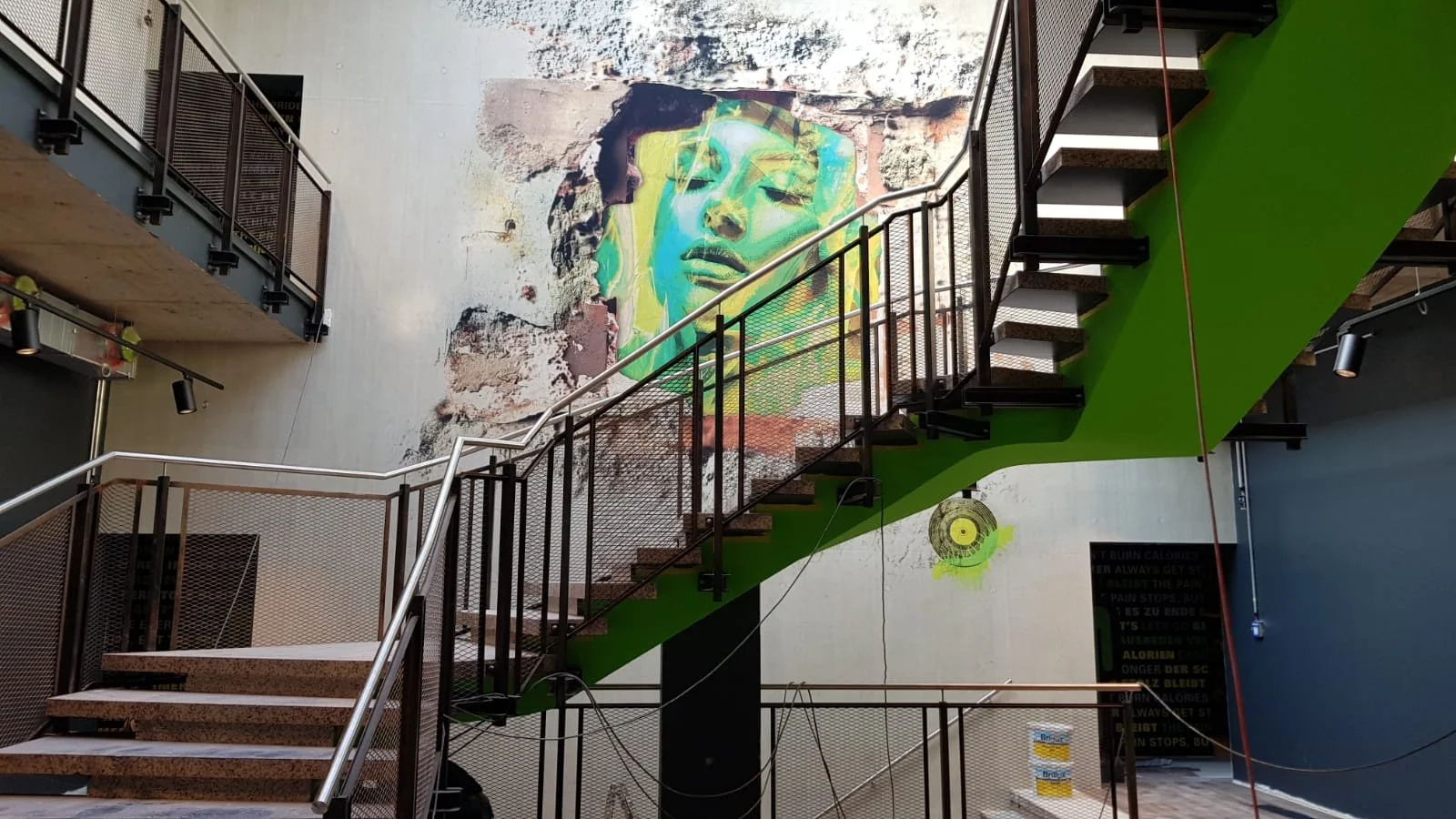 Innenraum eines modernen Gebäudes mit Wandmalerei eines stilisierten Gesichts, einer Treppe mit grüner Wandverkleidung und weiteren Kunstwerken an der Wand.