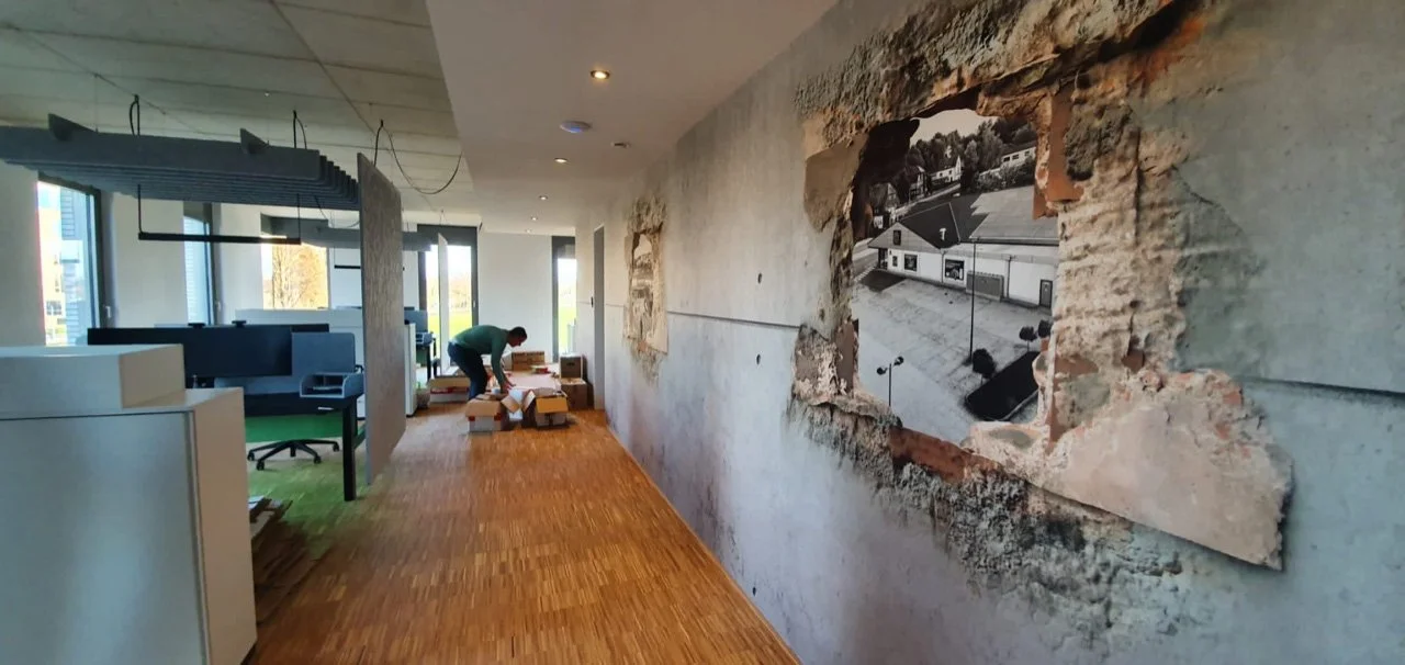 Mitarbeiter arbeitet an der Renovierung eines modernen Bürogebäudes, die Wand wird gerade neu gestaltet, sichtbare Holzboden, große Fenster, die viel natürliches Licht hereinlassen, und ein behutsam repariertes Wandstück mit einem alten Foto.