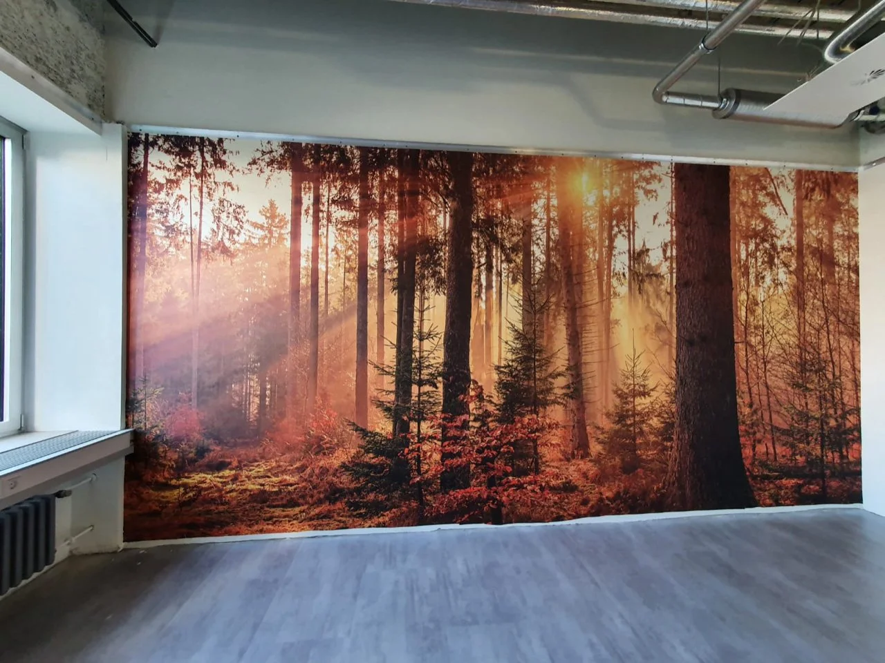 Wand mit gedrucktem Waldmotiv in warmen Orangetönen, simuliert Sonnenstrahlen durch Bäume, in einem Raum mit Fenster und Holzfußboden.