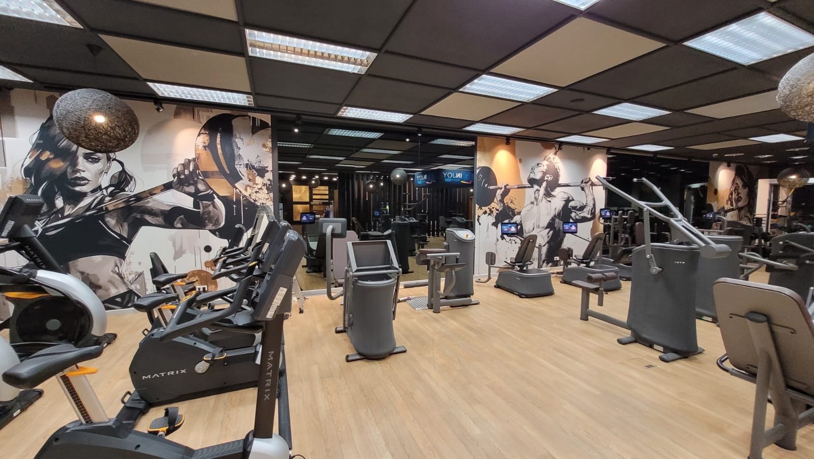 Fitnessstudio mit Cardio- und Krafttrainingsgeräten, Wandmalereien von Sportlern, helle Beleuchtung und Holzboden