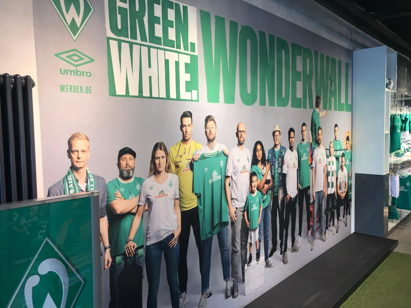 Eine Wand mit einer politischen Werbeaktion für den Fußballverein Werder Bremen, zeigt Menschen in Werder-Trikots und -Pullovern, some halten Trikots hoch, im Hintergrund groß geschriebene grüne Worte »GREEN. WHITE. WUNDERWAL.«