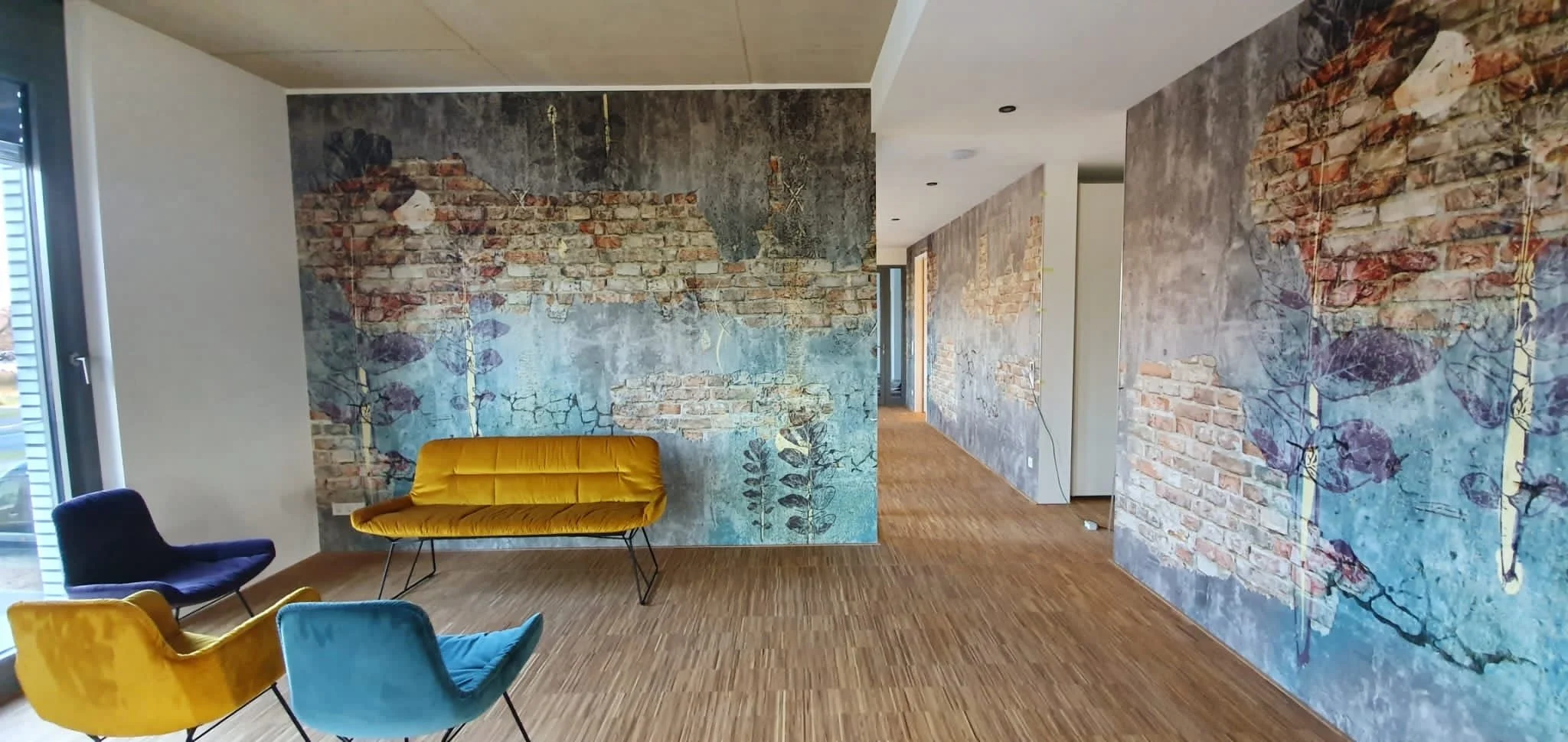 Innenaufnahme eines Gebäudes mit Wandmalerei, die eine verblasste Backsteinwand mit bläulichem, kollagenem Fresco-Effekt zeigt, und vier bunte Samtstühle in der Nähe einer großen Fenstertür.