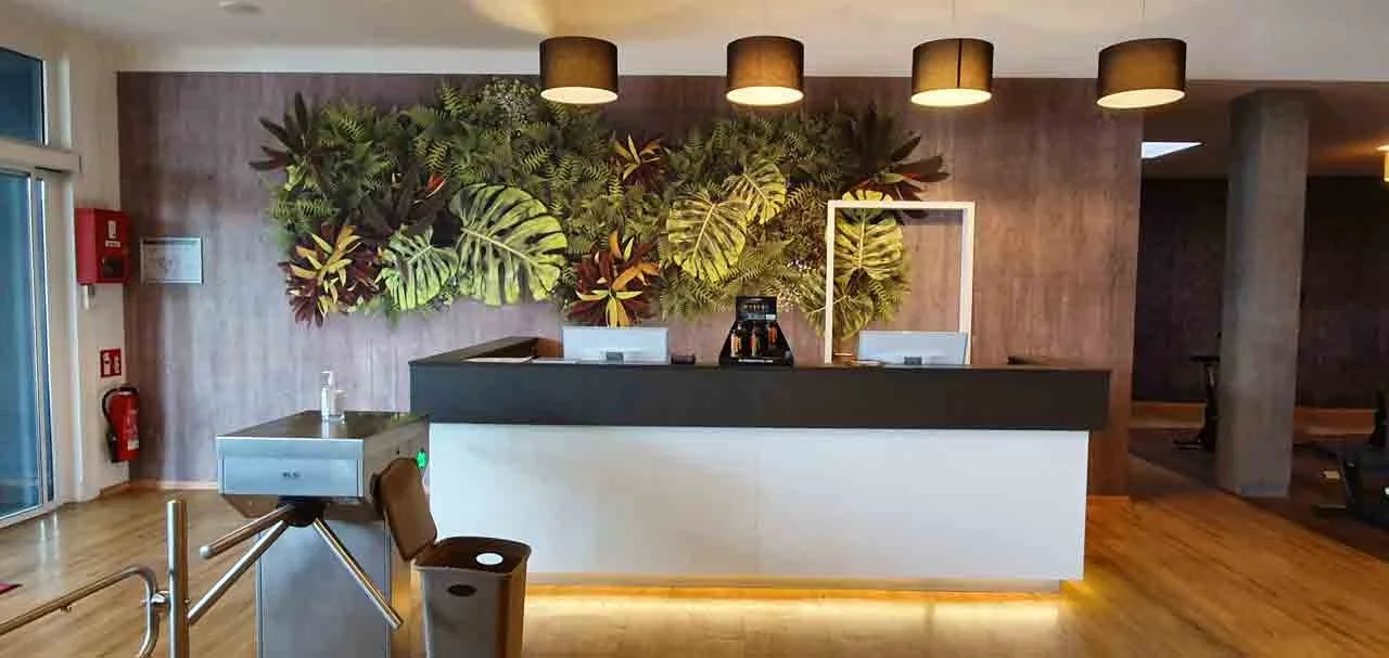 Rezeption im Eingangsbereich eines Hotels, mit einem dunklen Empfangstresen, zwei Computern, einer Kaffeemaschine und einer großen Wanddekoration aus grünen Blättern. Neben der Rezeption sind ein Handwaschbecken, ein Feuerlöscher, eine Wand mit Feuer