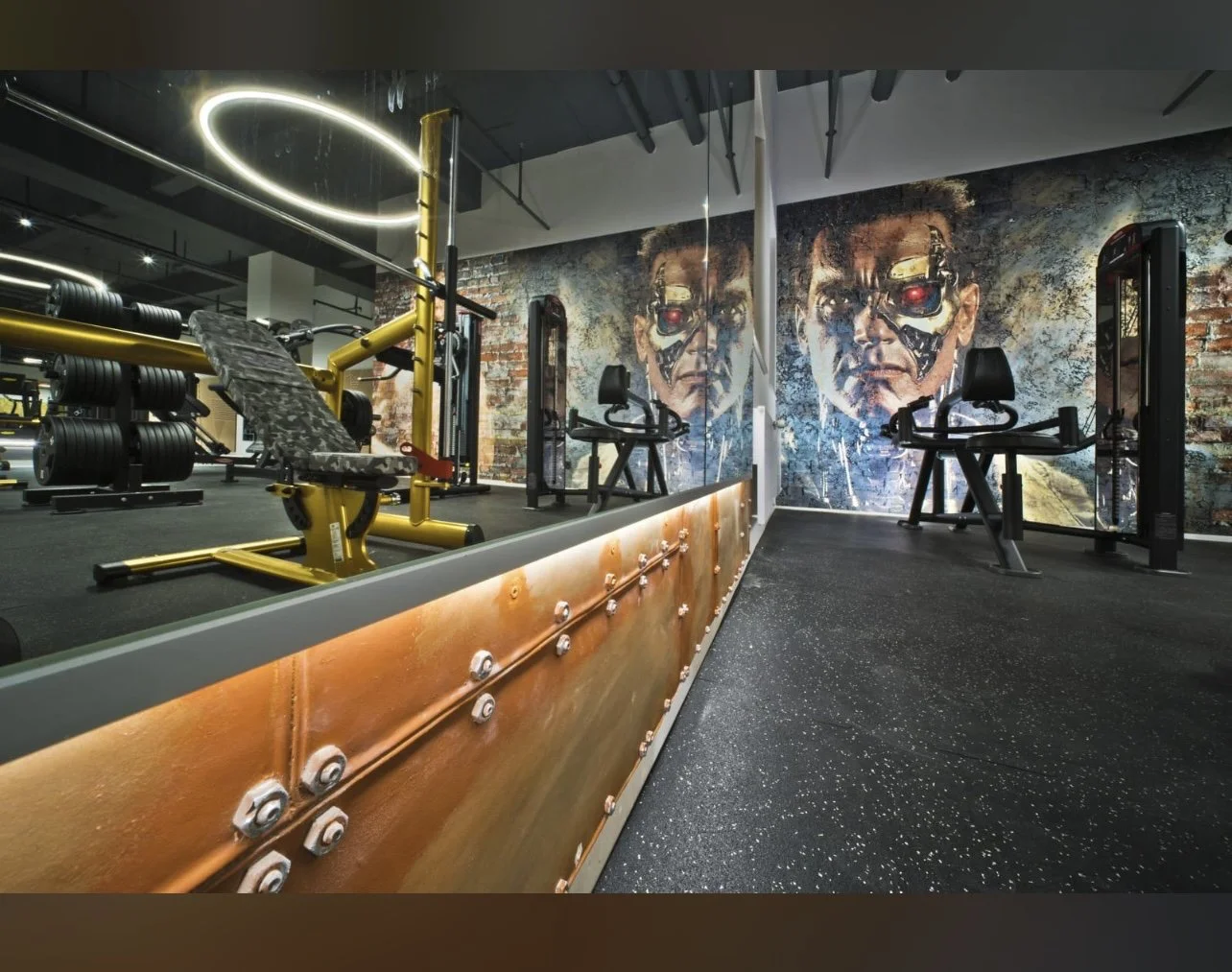 Fitnessstudio mit Krafttrainingsgeräten und Wandgemälde eines maskierten Mannes mit Cyborg-Elementen