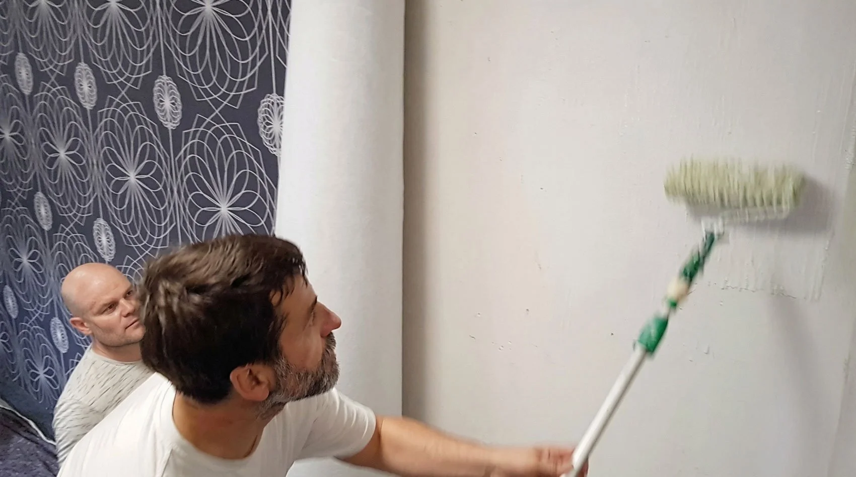 Zwei Männer streichen eine weiße Wand mit einer Farbrolle. Im Hintergrund ist eine dunkelblaue Vorhang mit weißen Mustern zu sehen.