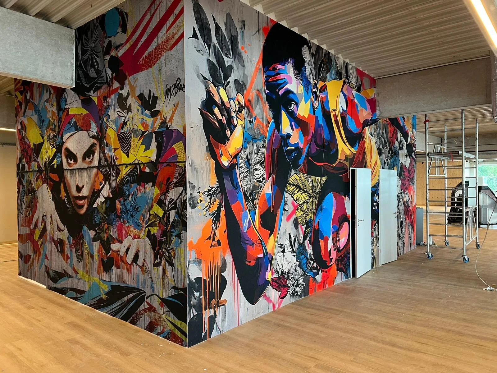 Graffiti-Malerei an einer Wand, die zwei Personen zeigt. Die linke Person ist eine Frau mit dunklen Haaren und intensivem Blick, die rechte Person ist ein Mann, der in die Kamera schaut und in einer kraftvollen Pose steht, umgeben von floralen und ab