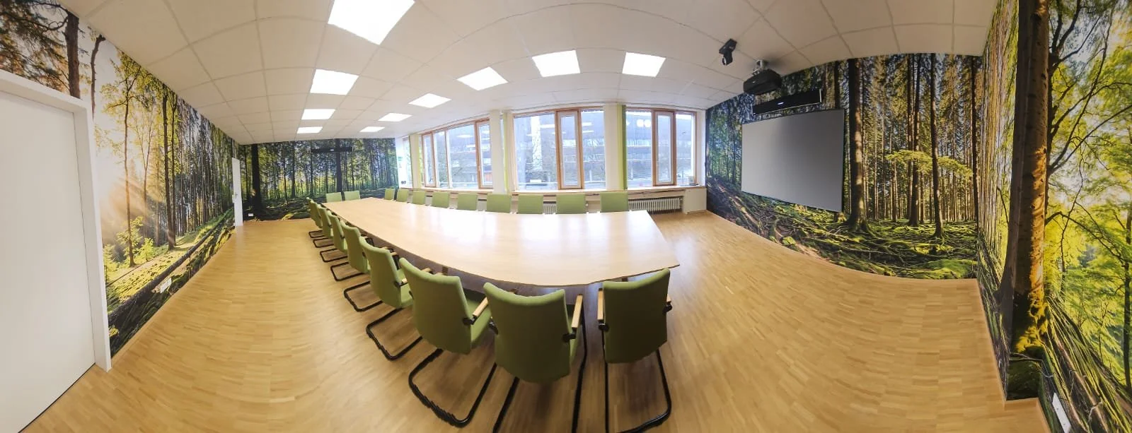 Moderner Konferenzraum mit großem rechteckigem Tisch, grünen Stühlen, Wandbemalung mit Waldmotiv, großen Fenstern und Leinwand auf der Wand.