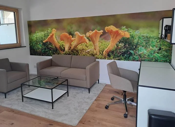 Wohnzimmer mit grauer Couch, einem Sessel, Glastisch, Holzfußboden und einer großen Wandbild mit Pilzen und Naturlandschaft.