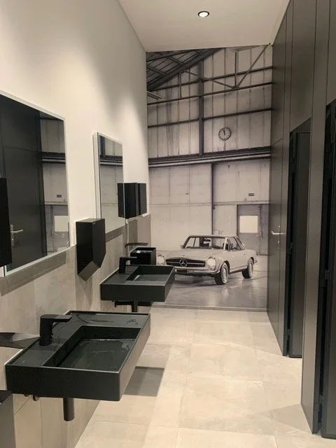 Ein Badezimmer mit schwarzen Waschbecken, Spiegeln und Handtuchspendern, im Hintergrund befindet sich ein Wandbild oder Druck mit einem klassischen Auto in einer Garage oder Werkstatt.