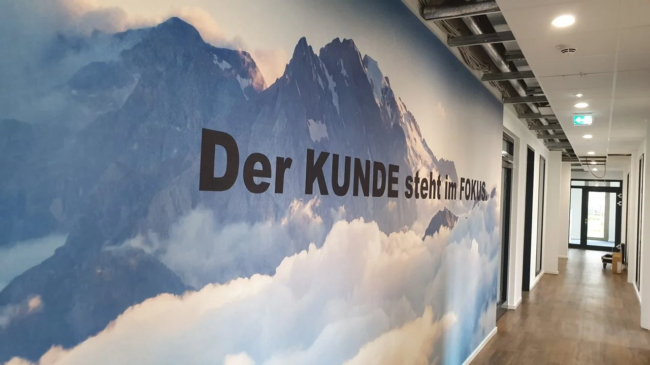 Büroflur mit Wandbild von Bergen und Text 'Der KUNDE steht im FOKUS'.