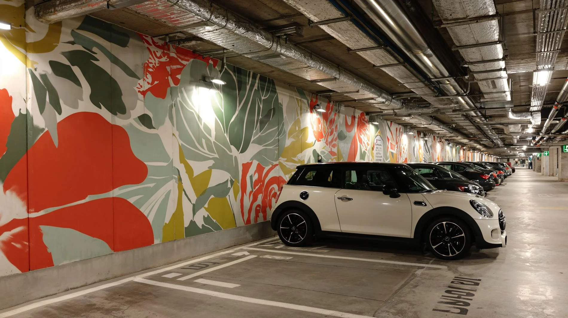 Parkhaus mit bunten Wanddekorationen, mehrere geparkte Autos, darunter ein weißer Mini Cooper im Vordergrund.