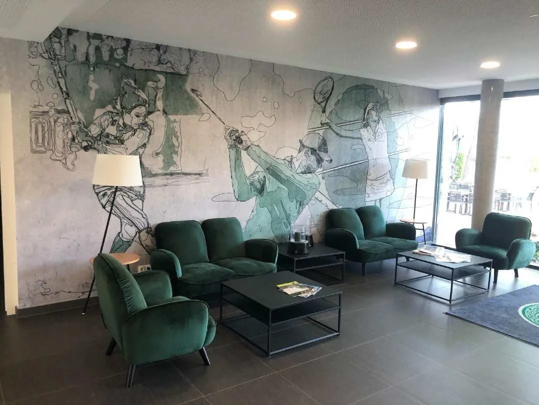 Innenraum eines Wartezimmers mit grünen Sesseln, Lampen, einem Tisch und einer Wand mit einer großen Wandmalerei von Tennisspielern.