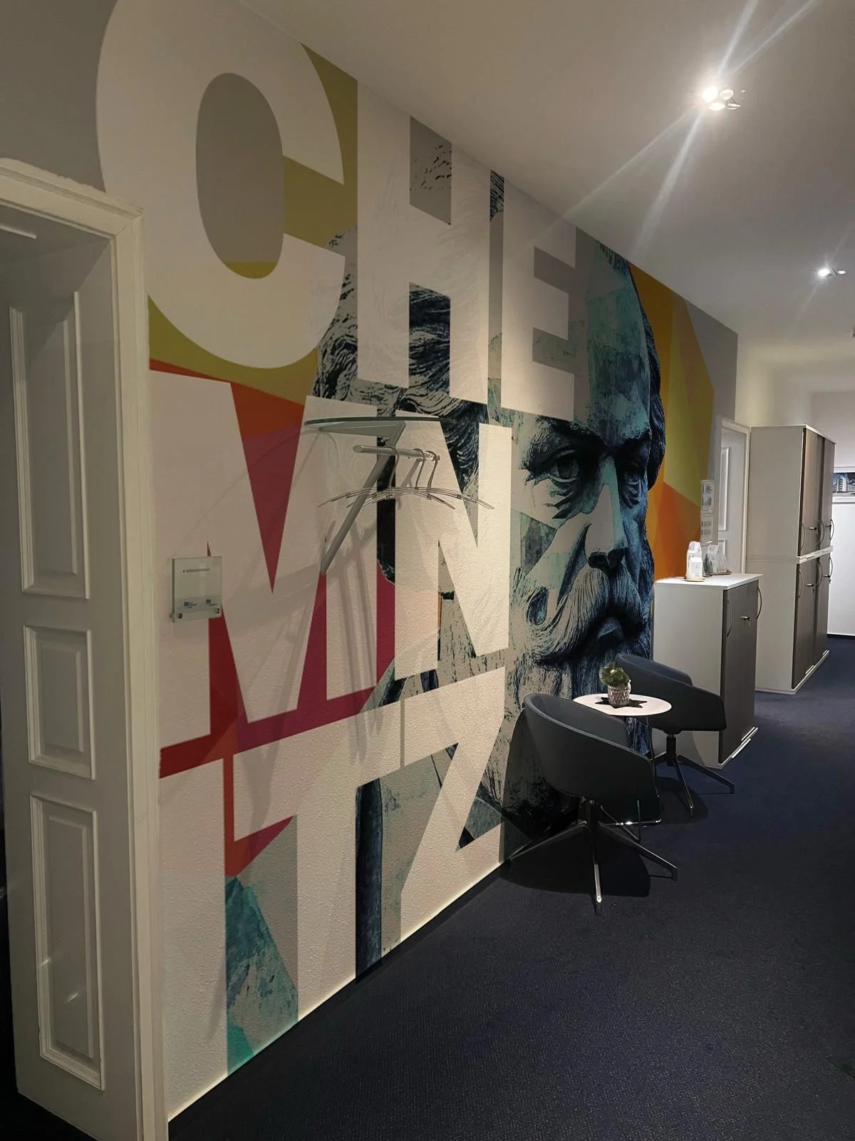 Wandtapete mit großem, stilisiertem Portrait eines Mannes mit Bart neben buntem geometrischem Design, im Raum mit Tür und Sitzgelegenheiten.