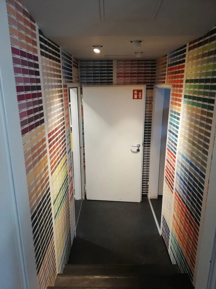 Kleines Flur oder Raum mit Wänden, die mit Farbmustern bedeckt sind, und einer weißen Tür mit einem Feuerlöscher-Schild darüber.