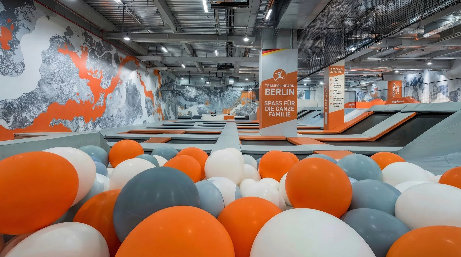 Innenansicht des Trampolinparks Berlin mit bunten Bällen, Trampolinen und Wanddekoration im Stil einer Weltkarte, orange und weiß dominierende Farben, mit Beschilderungen für Regeln und Sicherheit.