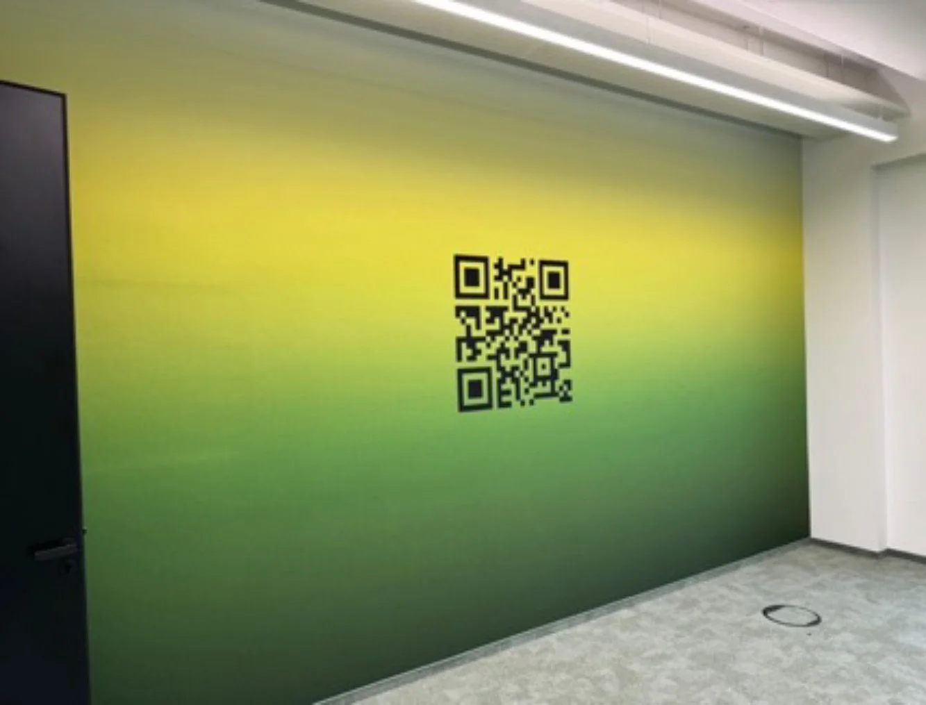 Ein grüner Wandabschnitt mit einem QR-Code in der Mitte, in einem modernen Raum mit grauem Boden und weißen Wänden.