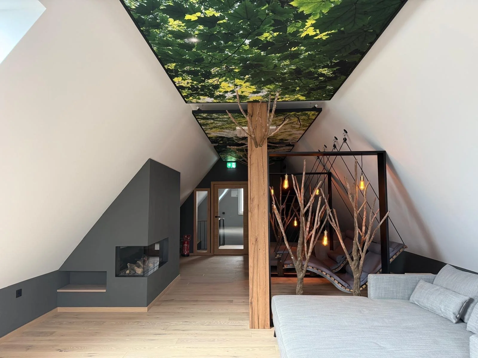 Ein modern eingerichteter Dachboden mit weißen Wänden, einem grauen Kamin, einem weißen Sofa, einem Baum mit Lichterketten, einer Hängesitz-Hängematte und einer Deckenabbildung von einem Wald mit grünem Laub.