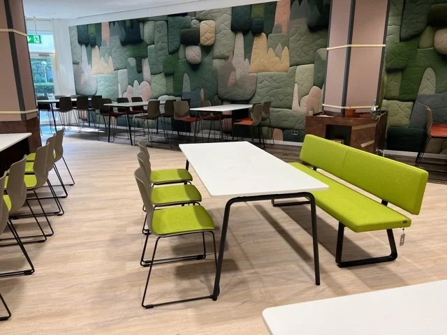 Ein moderner, leerer Aufenthaltsraum oder Gemeinschaftsraum mit Tischen, Stühlen und einer Wand mit Wanddekoration aus Polsterstücken in verschiedenen Grüntönen.