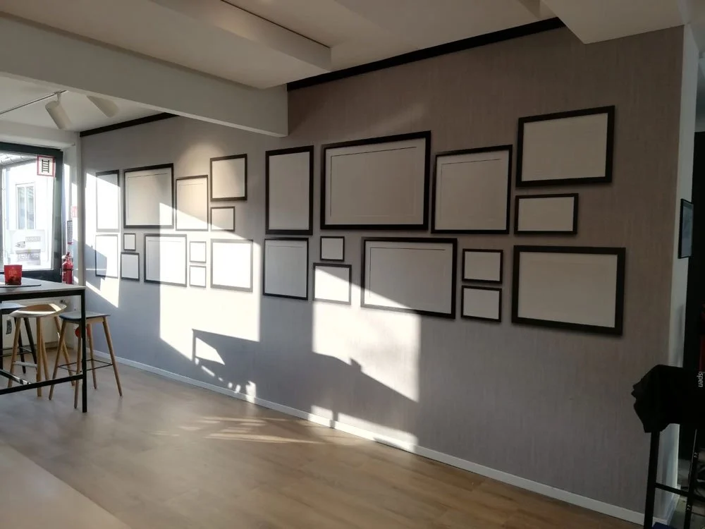 Leerer Raum mit einer Wand, die mit mehreren leeren Bilderrahmen in verschiedenen Größen dekoriert ist, sonniges Licht, Blick auf einen Fensterbereich mit Stühlen und einem Tisch.