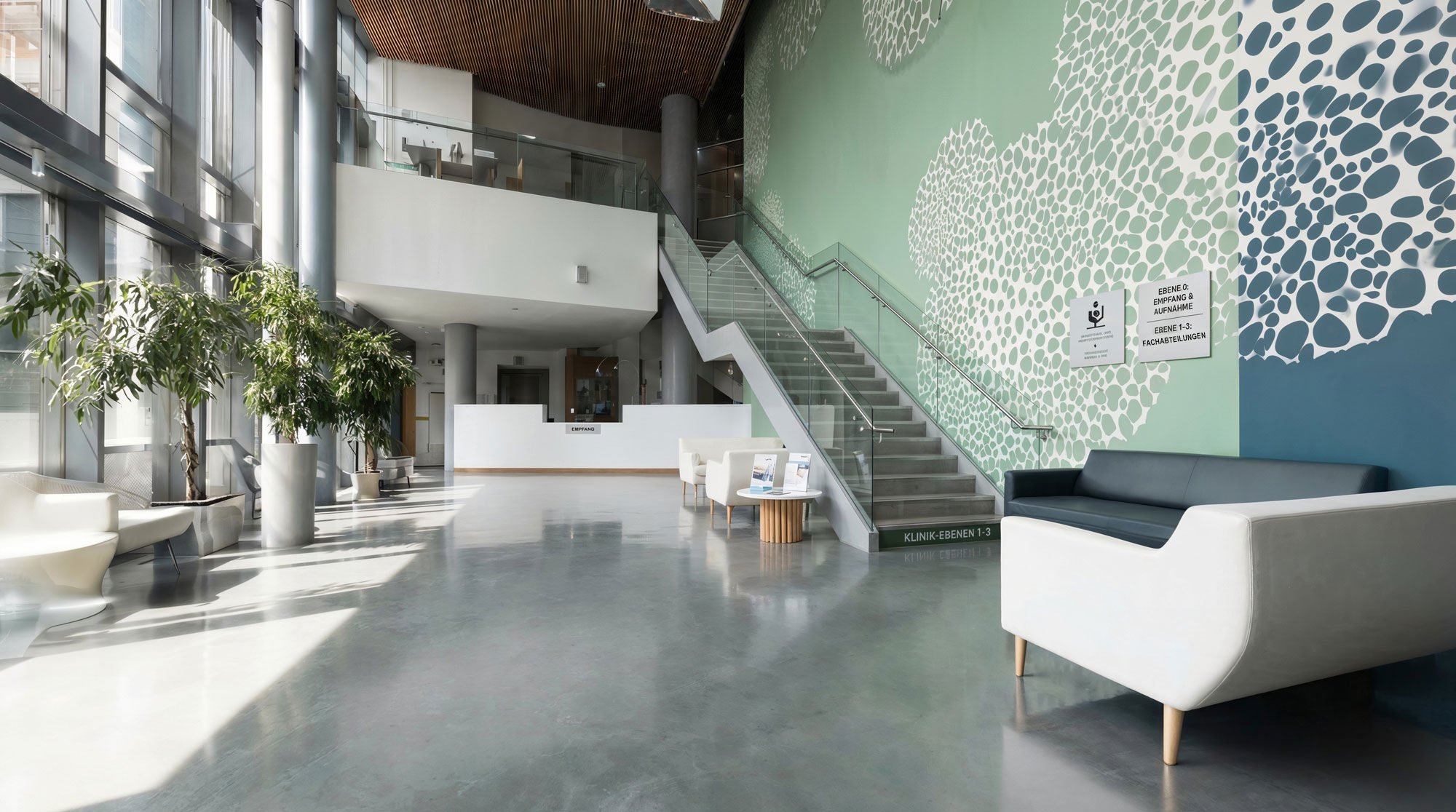 Heller, moderner Eingangsbereich eines Krankenhauses oder einer Klinik mit Sitzgelegenheiten, Pflanzen und einem Wandbild mit Baummustern.