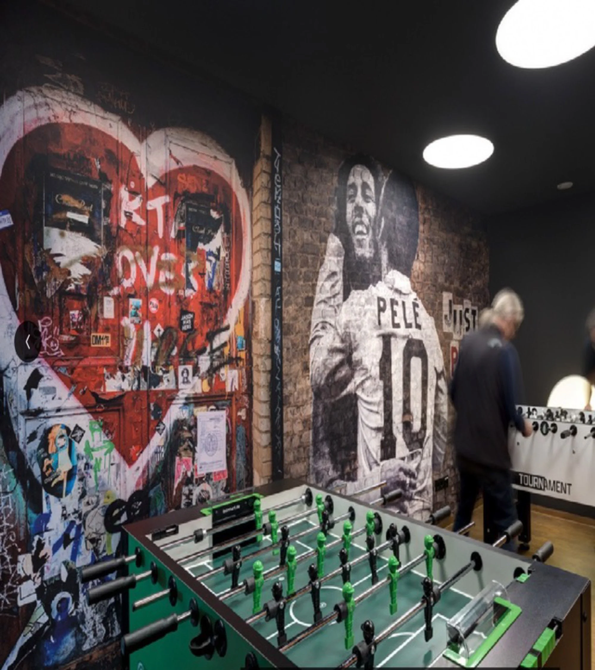 Ein Raum mit Street Art Murals an den Wänden, einem Tischfußballspiel und einigen Personen im Hintergrund.