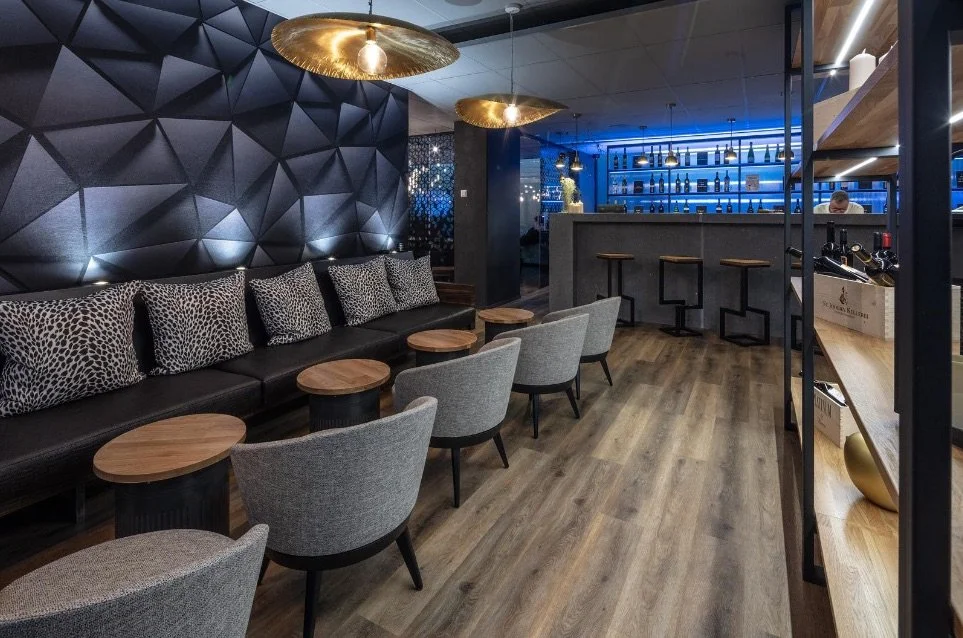 Ein modern eingerichteter Lounge- oder Barbereich mit schwarzen Wandanstrich, goldenen Deckenleuchten, einer schwarzen Ledercouch mit Leopardenmuster-Kissen, mehreren bequemen Stühlen und Tischen aus Holz, sowie einer Bar im Hintergrund mit blauer Be