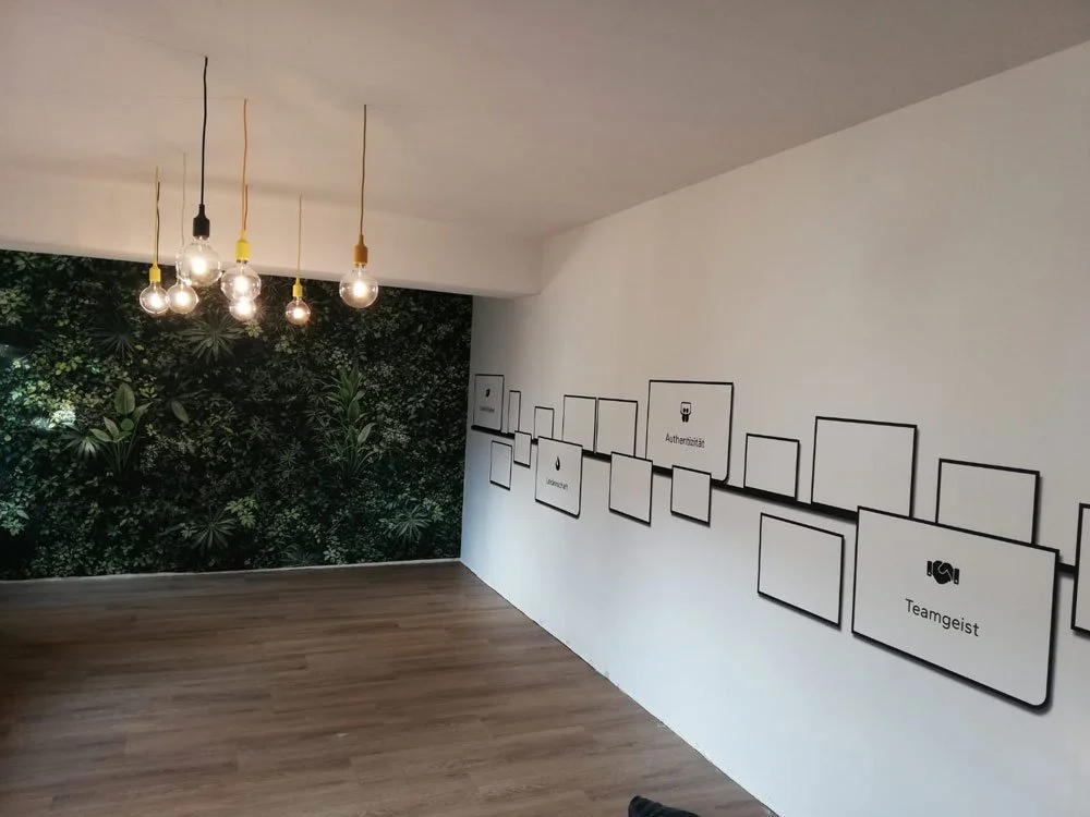 Ein leerer Raum mit Holzfußboden, einer Wand mit grüner Pflanzenmuster, und einer weißen Wand, die mit schwarzen Rahmen und Symbolen für Teamgeist, Authentizität und andere Begriffe dekoriert ist. Es hängen mehrere Glühbirnen an Kabeln von der Decke.