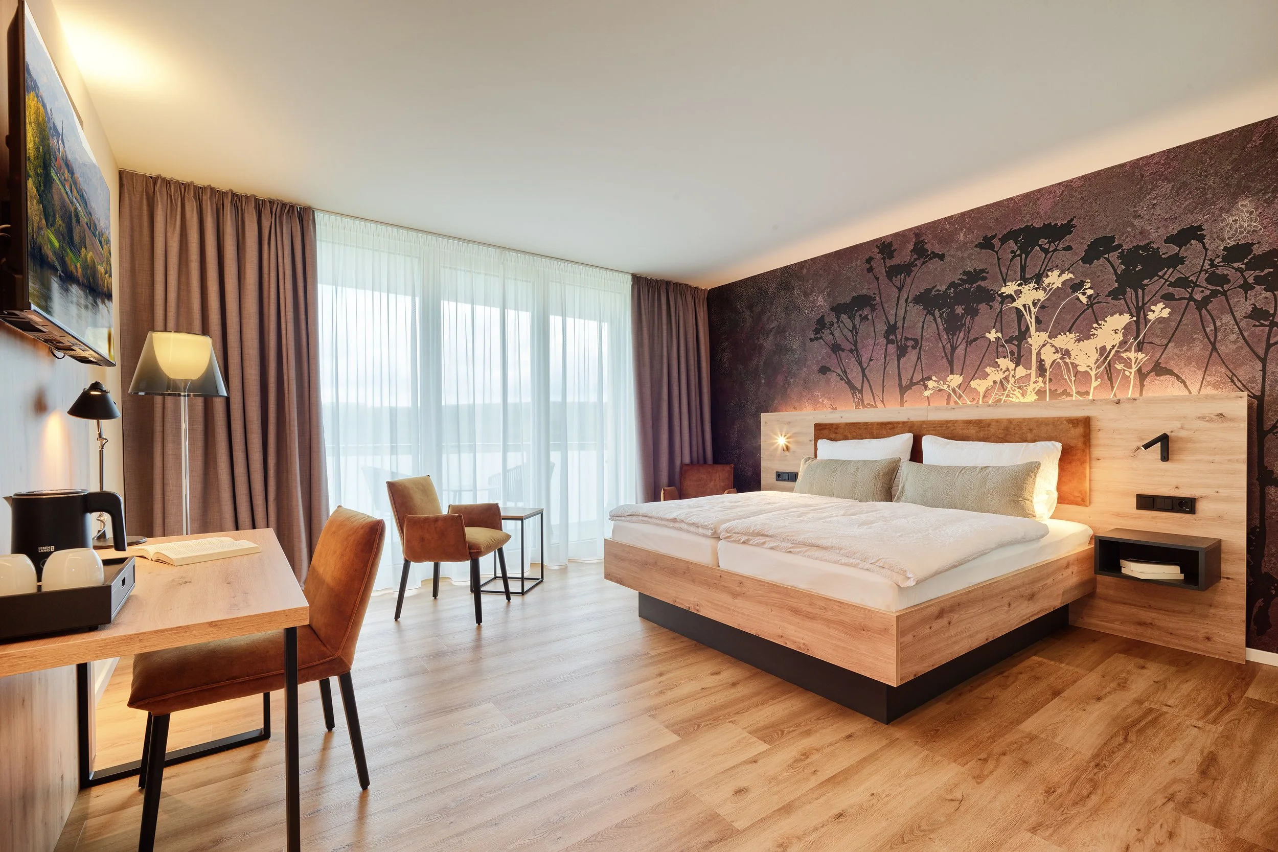 Ein modernes Hotelzimmer mit einem großen Bett, einem Wandbild mit Baum-Motiv, einem Schreibtisch mit Stühlen, einem Fernseher, einem Wasserkocher und großflächigen Fenstern mit Vorhängen.