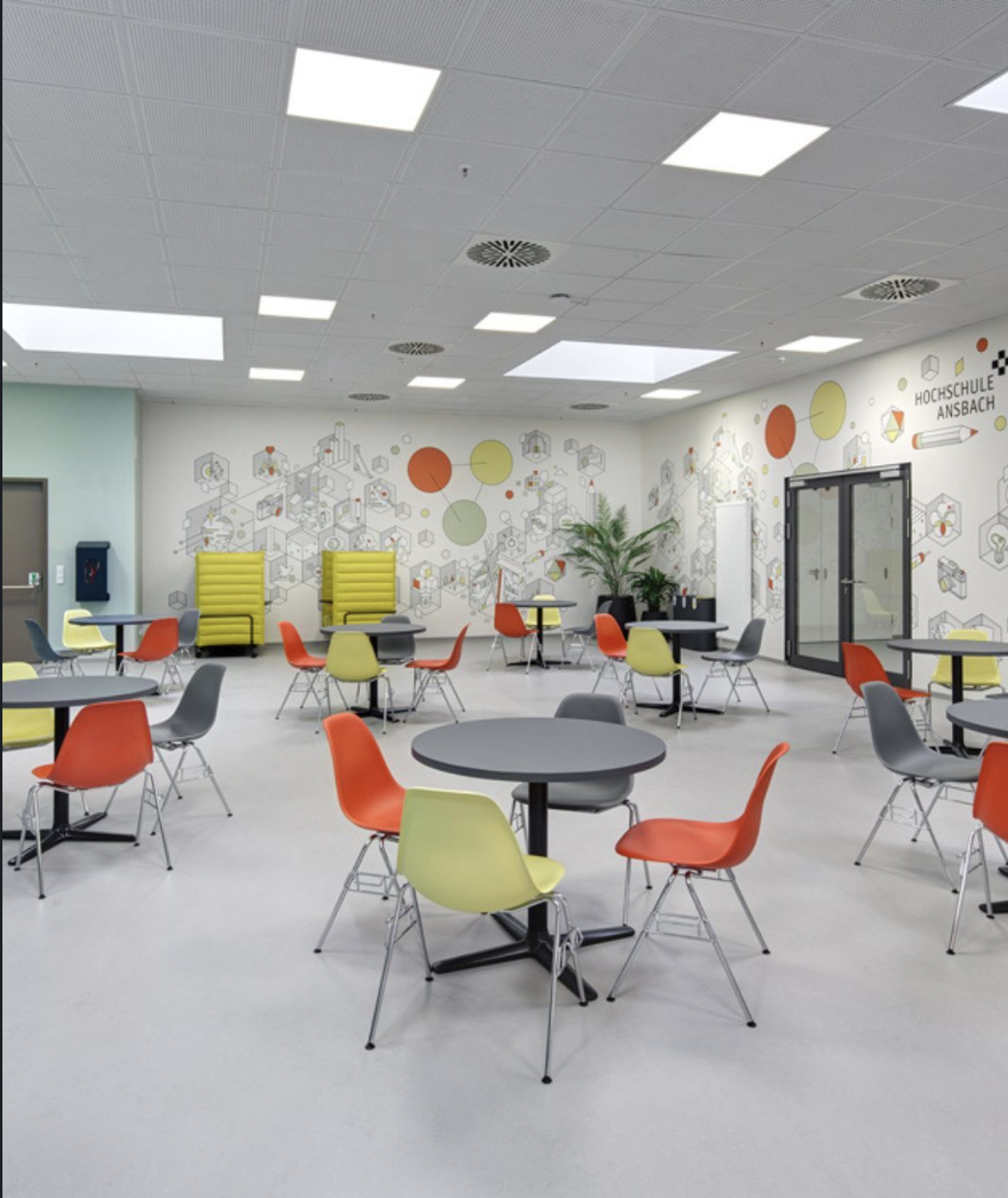 Ein heller Schul- oder Universitätsraum mit bunten Stühlen und runden Tischen, Wanddekoration mit geometrischen Mustern und der Aufschrift 'Hochschule Ansbach'.