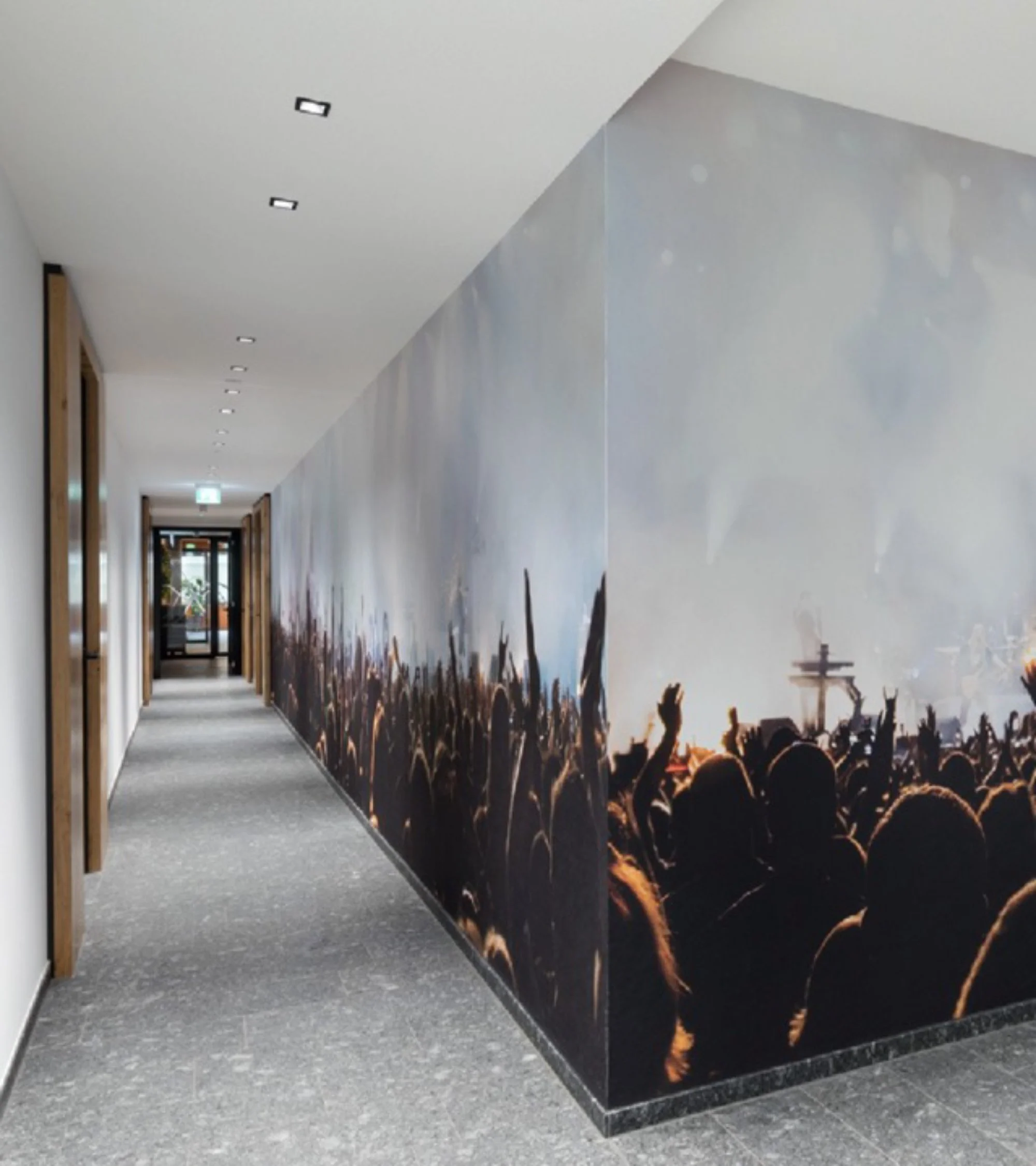 Innenansicht eines Flurs mit grauem Boden und weißen Wänden, eine seitliche Wand mit Wandbild eines Konzertpublikums bei Sonnenuntergang